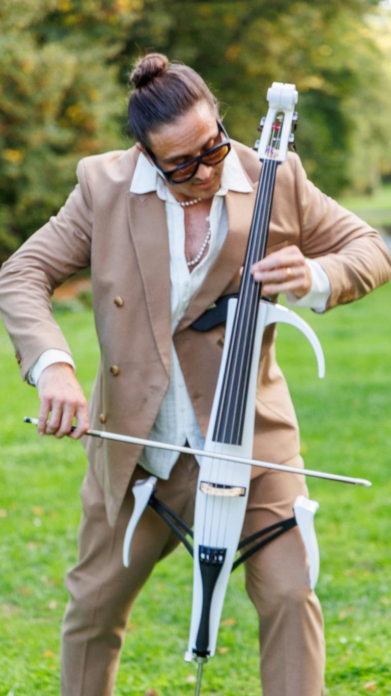 eleganter Live Musiker für besondere Anlässe Mann in weißem Anzug spielt ein Cello in einer parkähnlichen Umgebung.