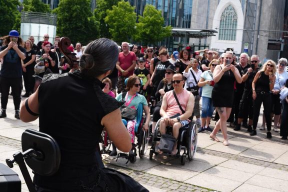 E-Cello beim WGT in Leipzig Eine Gruppe von Menschen in Schwarz beobachtet eine Darbietung, einige im Rollstuhl.